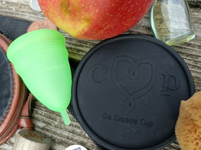 Sfeerplaatje van een groene menstruatiecup met een appel, een zwarte sterilisator en een sponsje liggend op een vlonder